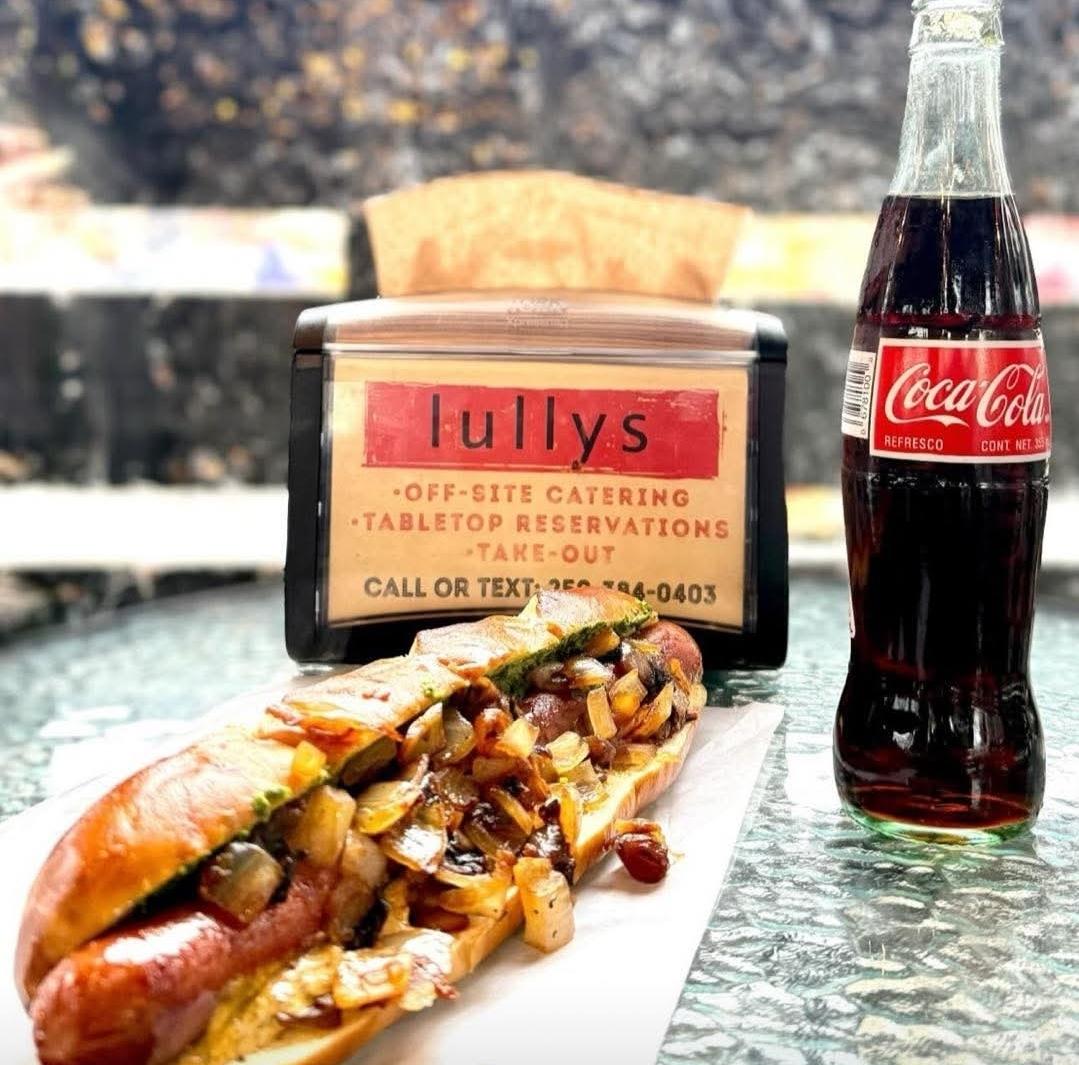 A Lullys hot dog on a pretzel bun with onions, next to a Coke and a Lullys take-out sign.